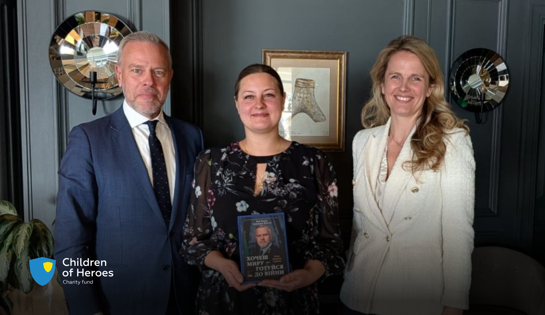 A representative of the Children of Heroes Charity Foundation holds a Ukrainian-language book, “If You Want Peace, Prepare for War,” standing between a man in a navy suit and a woman in a cream jacket in a dark interior with mirrored wall decor.